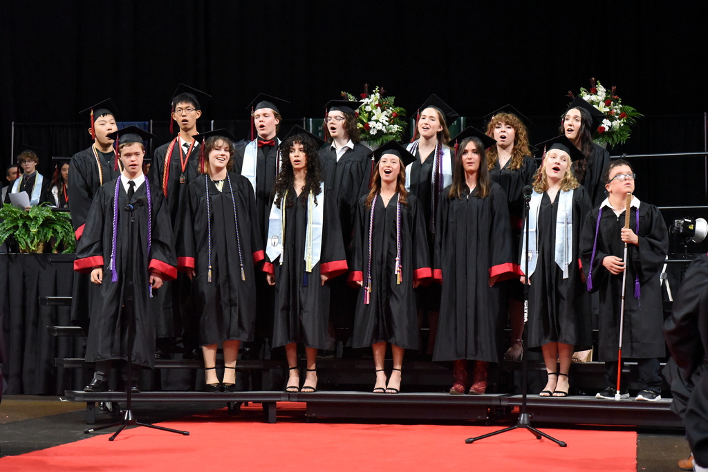 A group of students in black graduation robes singing on a stage with red carpet.