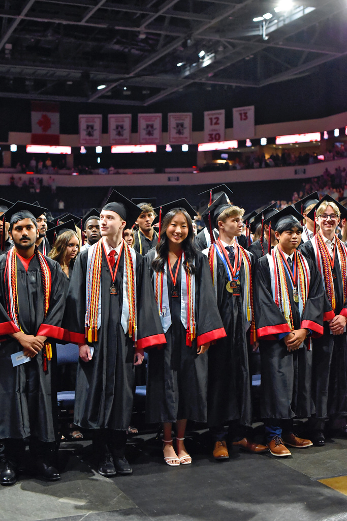 Group of graduates in academic robes and caps stand in line inside a stadium, with some wearing medals.