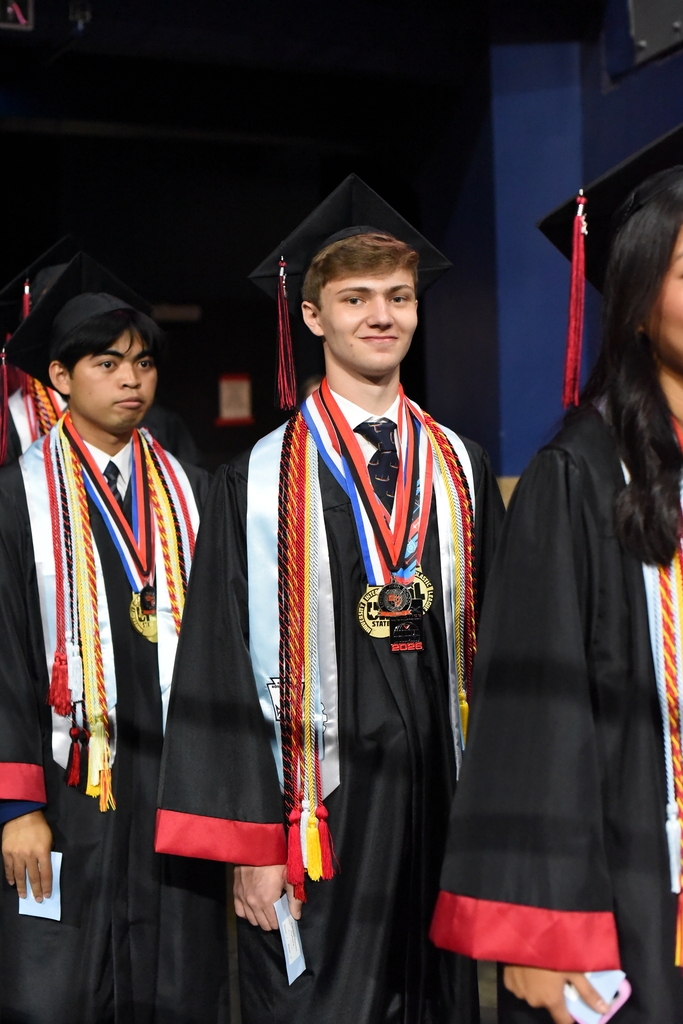 Four individuals in black academic gowns and hats stand in a row, facing forward. They wear medals with colorful ribbons.