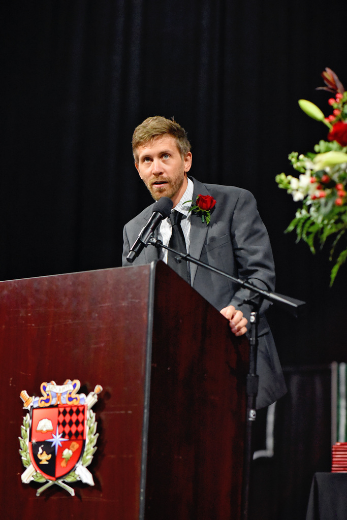 A man in a suit and tie speaks at a podium with a microphone. A red rose is on his lapel.