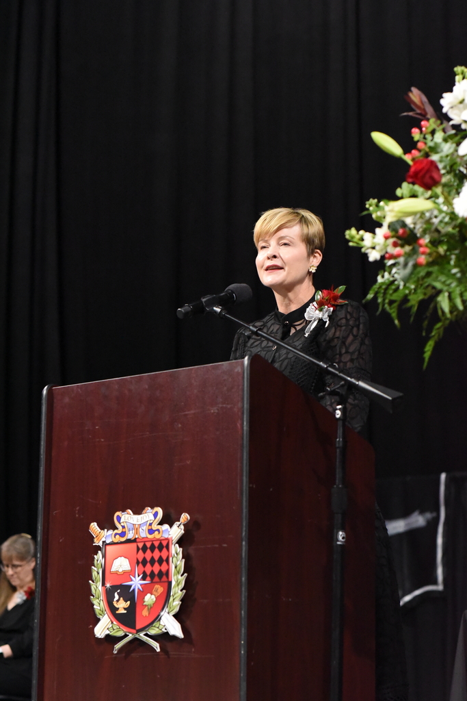 A woman speaks at a podium with a microphone. The podium features a shield-like emblem. A flower arrangement sits nearby.