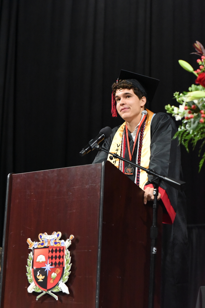 Person in graduation gown stands at a podium, speaking into a microphone, with a logo and flowers behind.