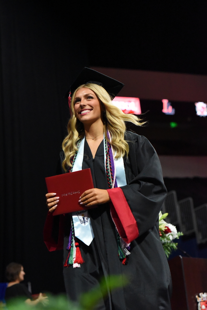 Woman with blonde hair wearing black graduation gown and cap holds a red folder with both hands, smiling.