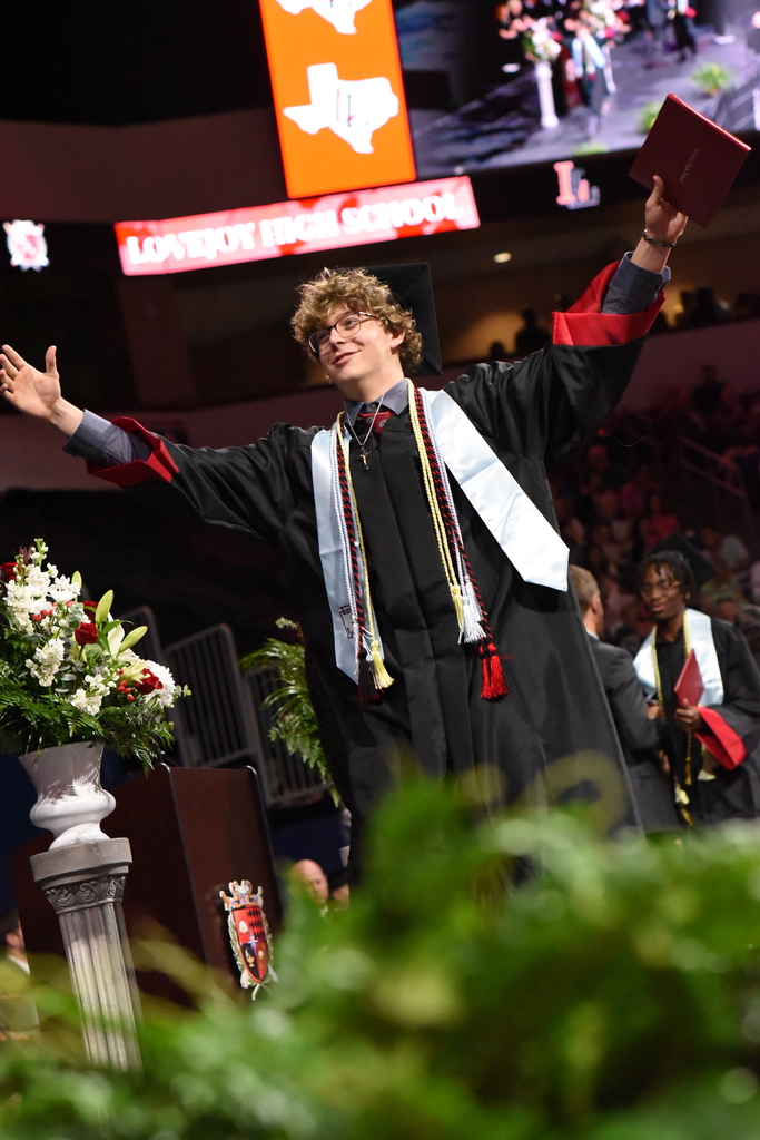 Person in graduation gown holds diploma high. Behind them, a podium with flowers and a screen displays a Texas flag.