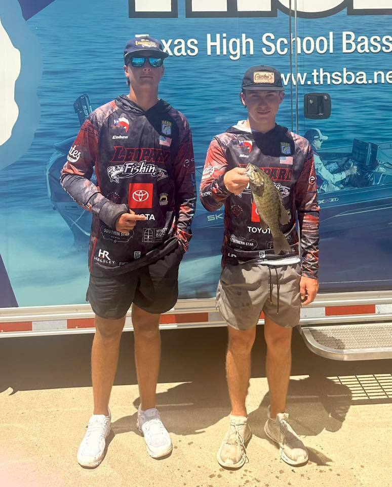 Two men in matching fishing gear pose with a small fish, standing in front of a banner advertising Texas High School Bass Association.