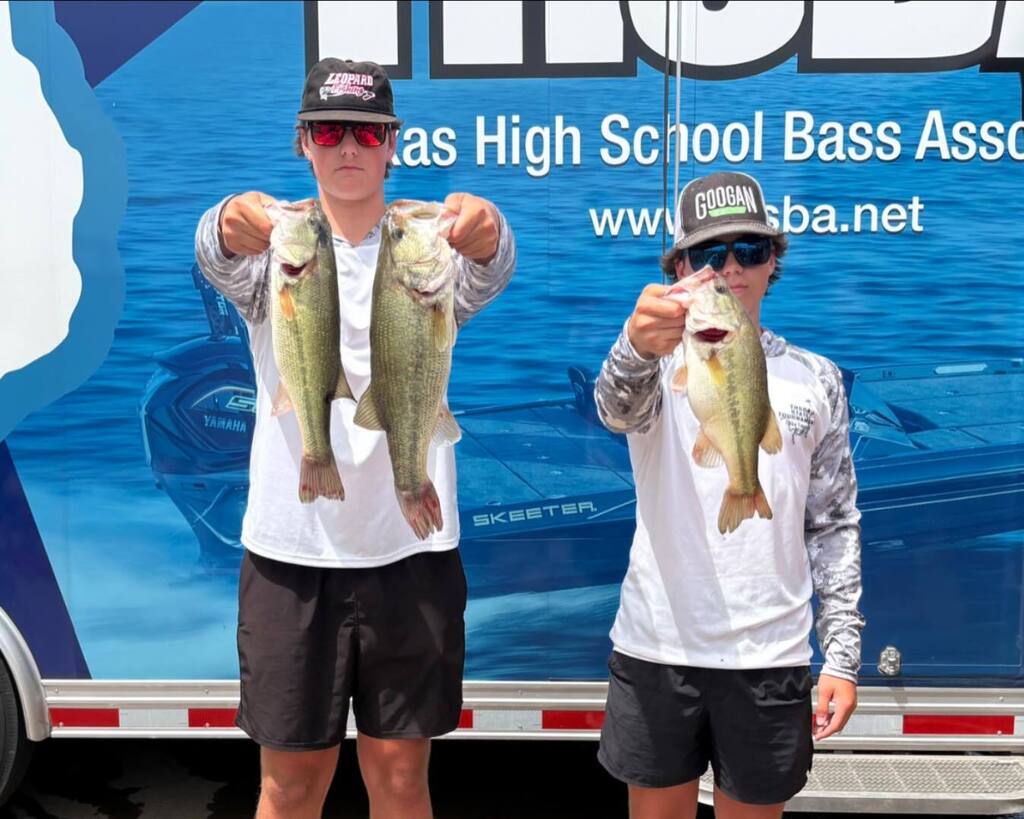 Two men hold up fish in front of a blue sign with text. Both wear white shirts, black shorts, and caps.