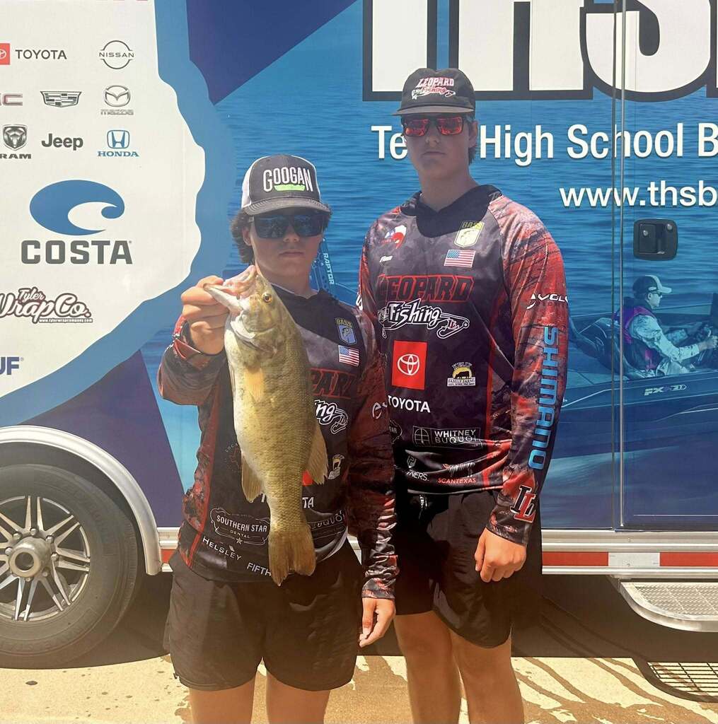 Two people stand by a vehicle, one holding a fish, both in fishing gear, with a backdrop of a Toyota Jeep banner.