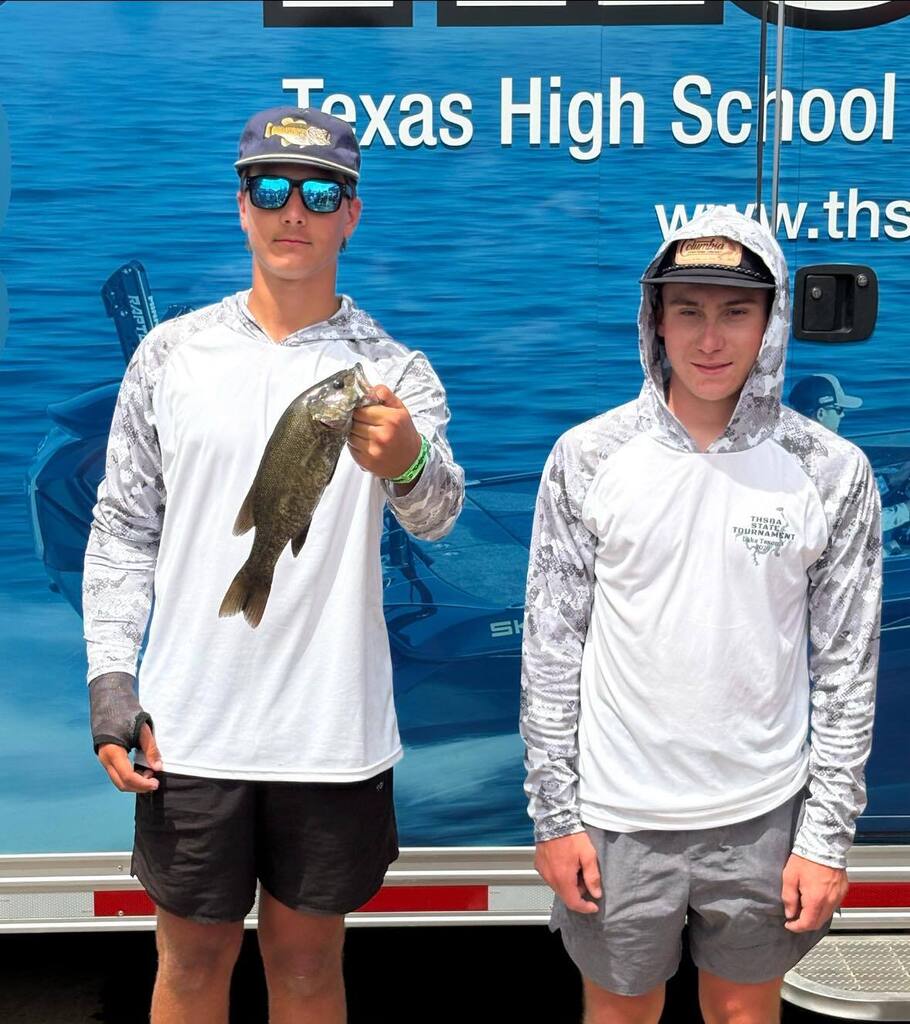Two people in matching shirts stand near a trailer. One holds a small fish, both wearing hats and sunglasses. A large, blue backdrop displays "Texas High School."