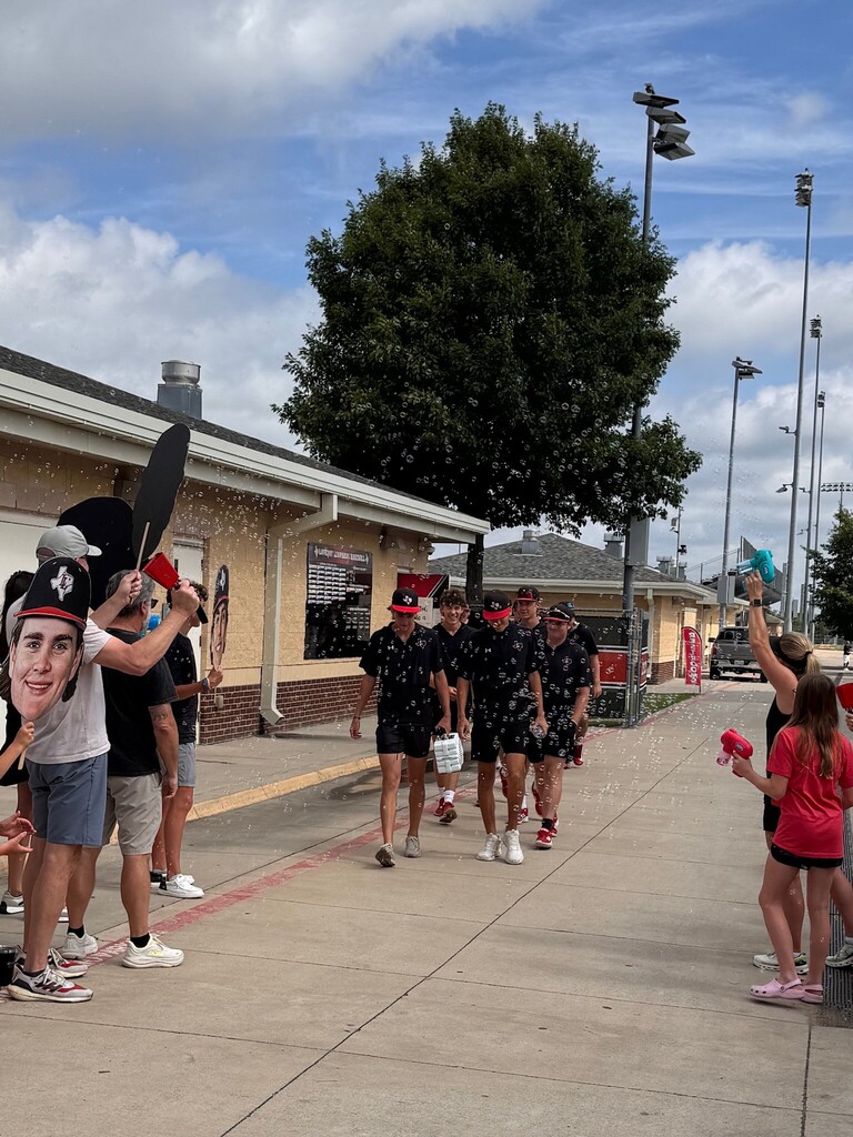 People in black uniforms walk on a sidewalk, with some holding water balloons. Trees and streetlights in the background.