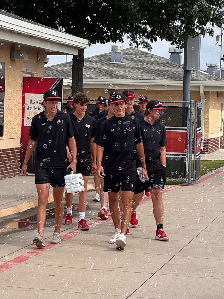 A group of people in matching black uniforms walk down a sidewalk, some holding bubble wands. A brick building and a fence are in the background.