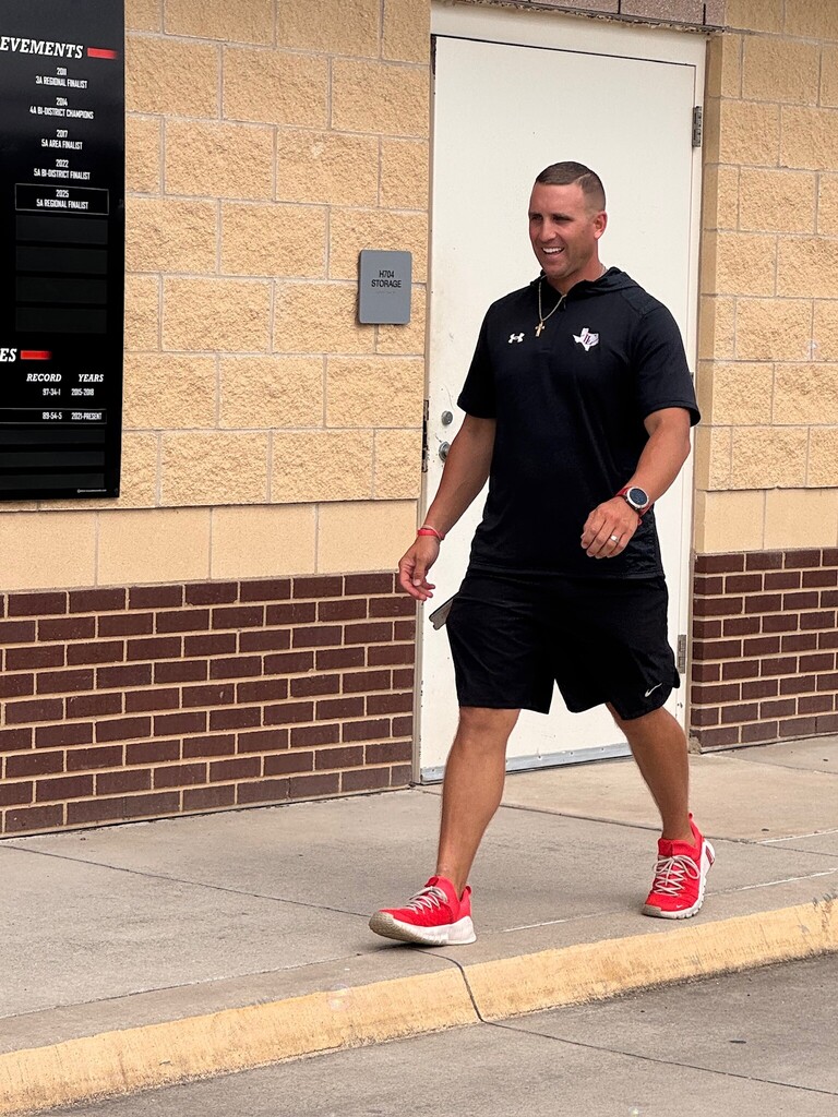 A man in black athletic wear and red sneakers walks along a sidewalk, near a building with a brick wall and a white door.