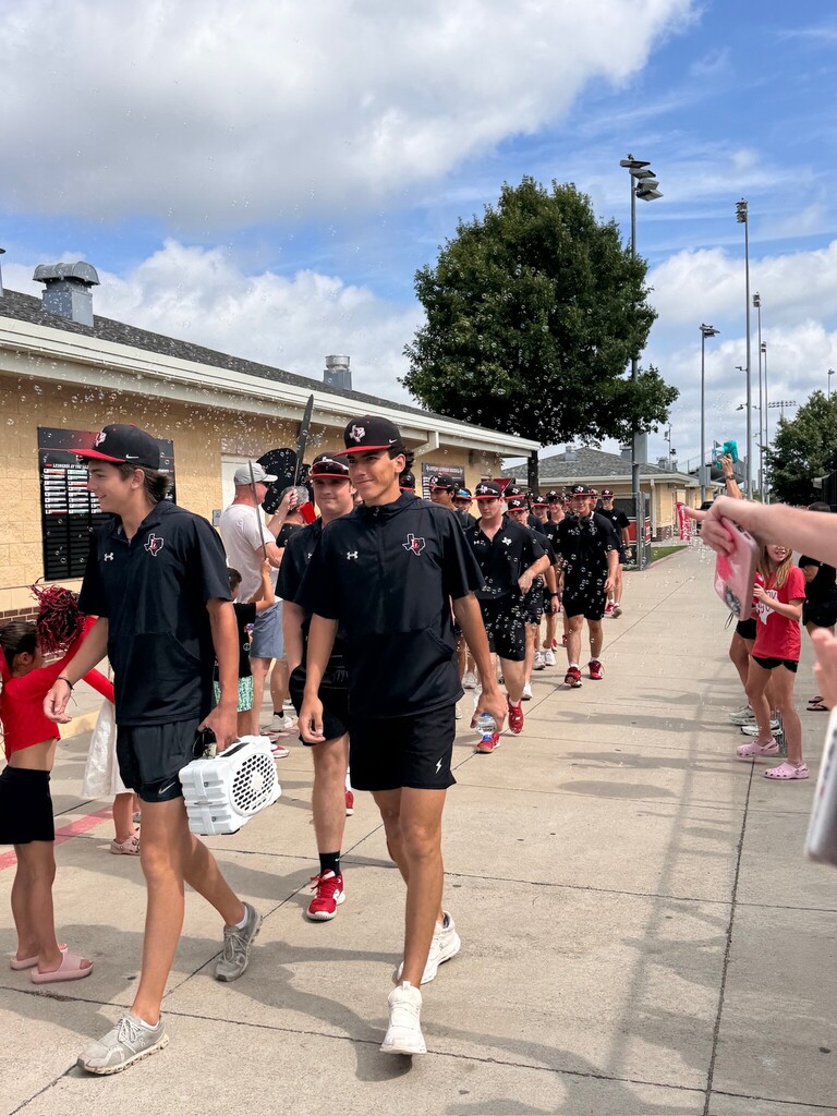 Group of people in matching black uniforms and caps walking down a sidewalk, one carrying a cooler.