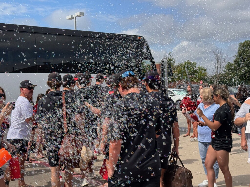 People celebrate in front of a bus with confetti; some wear hats and carry bags. Trees are in the background.