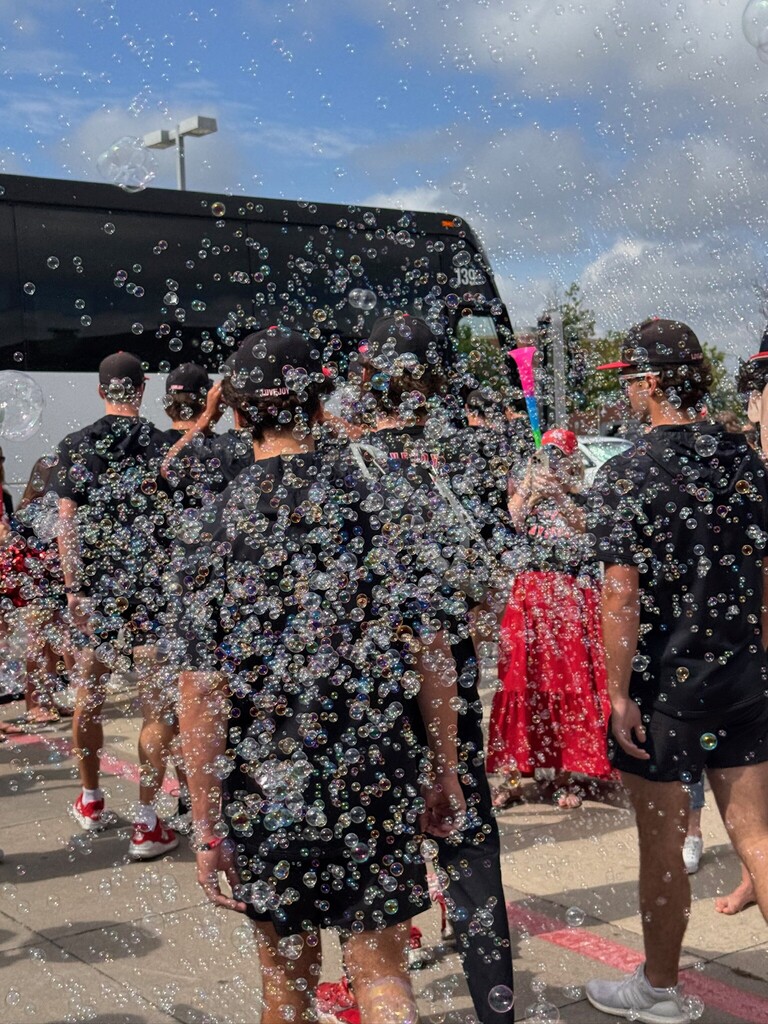 People in front of a bus under a blue sky. Bubbles float in the air. Some wear black clothes and sneakers.