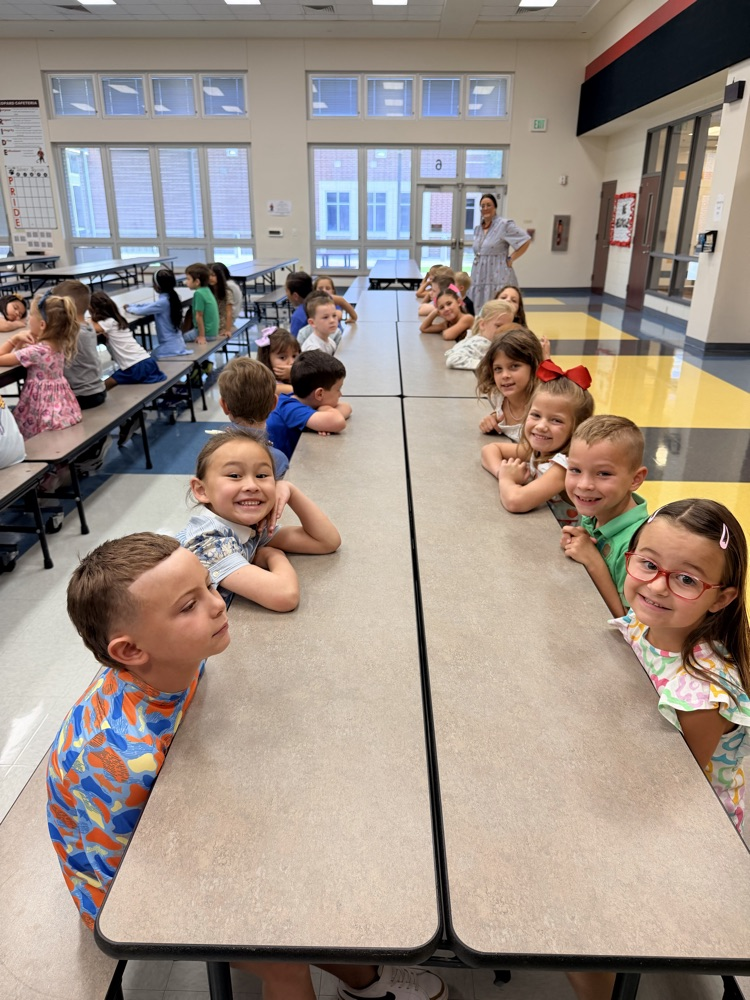 Kids in a classroom, seated at tables, some leaning forward to look over, with a teacher in the background.