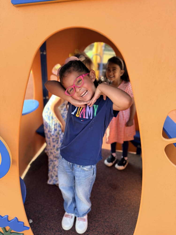 Child in blue shirt and glasses stands in an orange playhouse, smiling. Two girls, one in pink, stand behind.