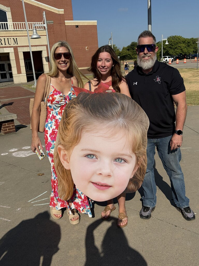 Three adults and a cutout of a girl's head with blonde hair and blue eyes. They stand outside a building.