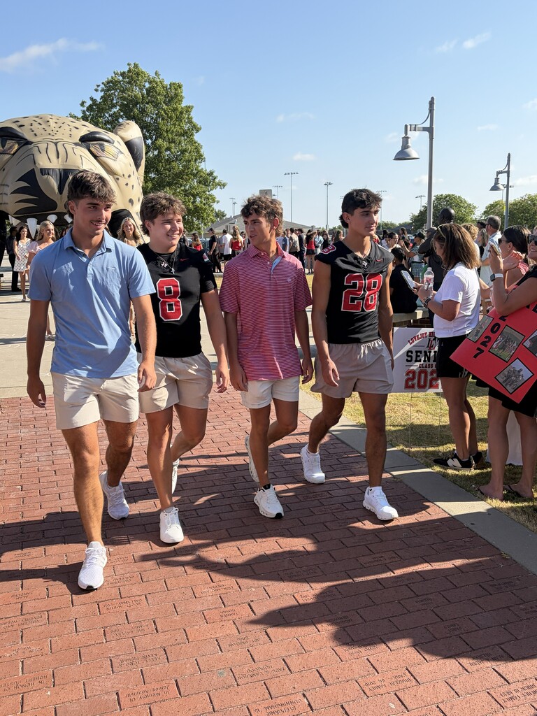 Four males walk together on a brick path, wearing athletic uniforms with numbers. Behind them, a large cat statue stands.