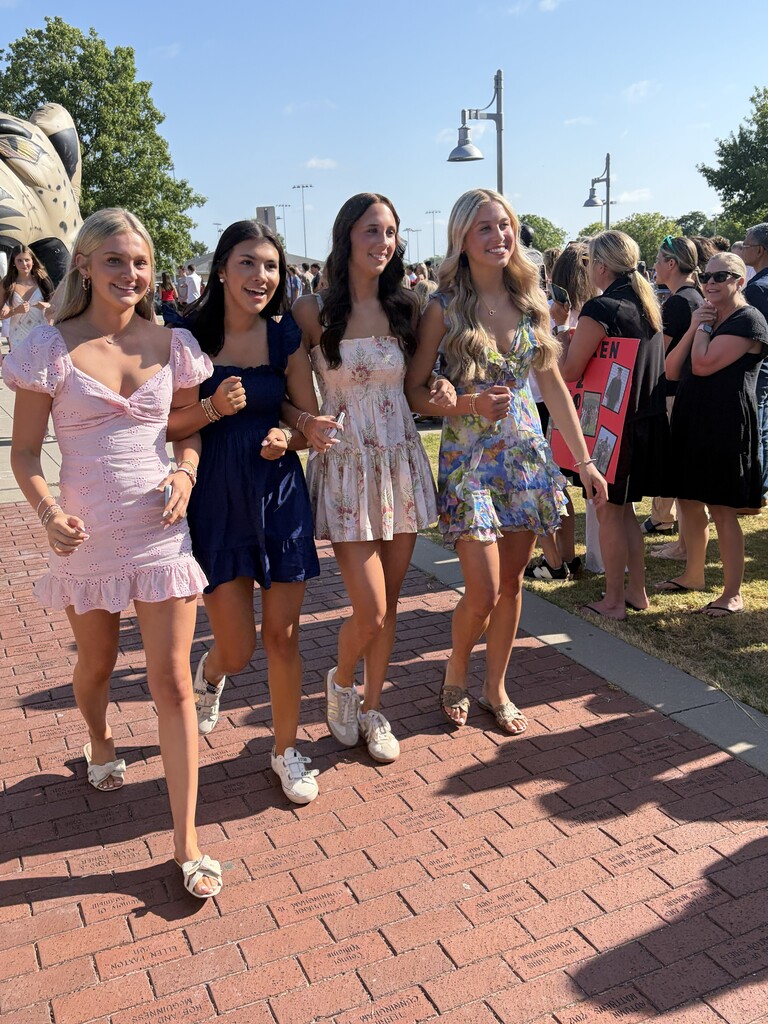 Four women walk on a brick path. They wear dresses of varying colors. Behind them, a large statue is visible.