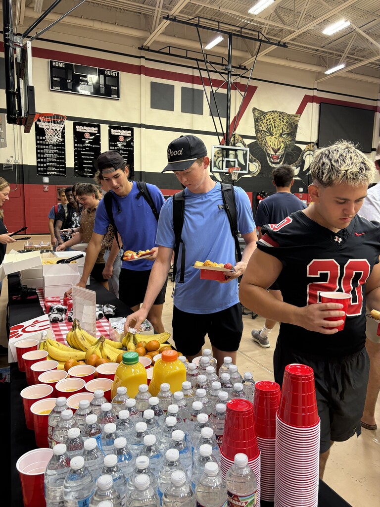 People gather around a table with food, drinks, and supplies at an event, possibly a sports gathering.