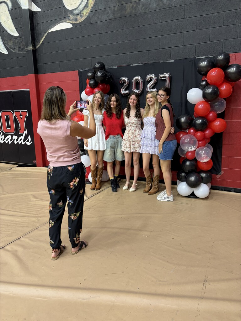 Five girls in summer clothing pose for a photo while a woman behind them takes a picture.
