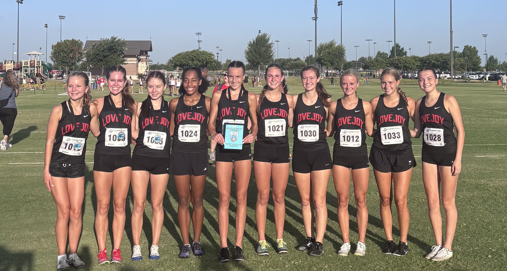 Seven female athletes stand in two rows, smiling. They wear black uniforms with white numbers. A trophy is in the middle.