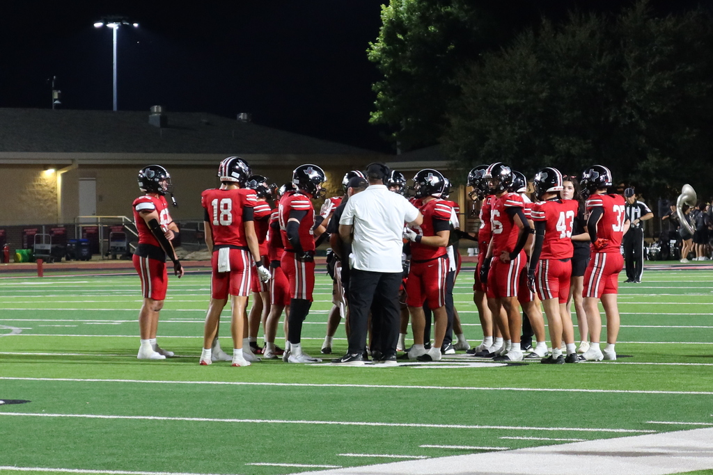 Team in red uniforms with numbers 18, 6, 42, and 3 huddles with coach on a field at night.