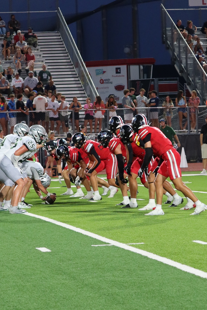 Football players in red and gray uniforms line up on a field, with a crowd in the background.