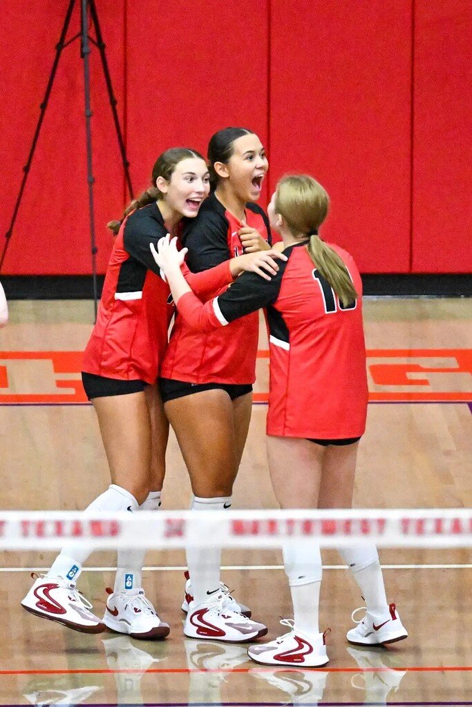 Four female volleyball players in red uniforms celebrate a point in a gymnasium with a red wall.