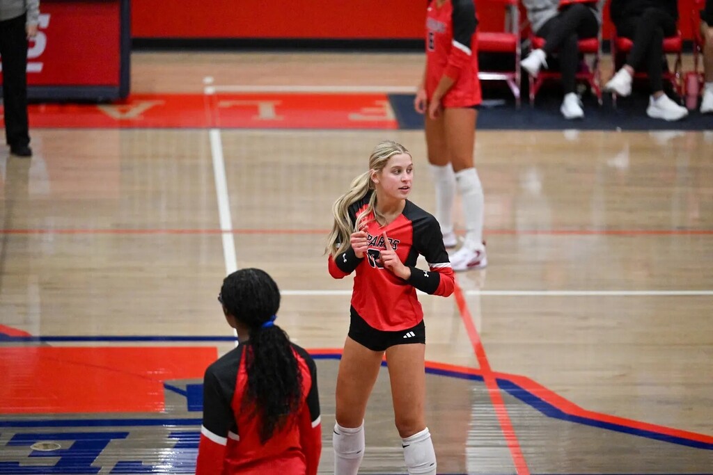 Volleyball players on a court. One player in red and black speaks while another listens. Red and white lines mark the court.