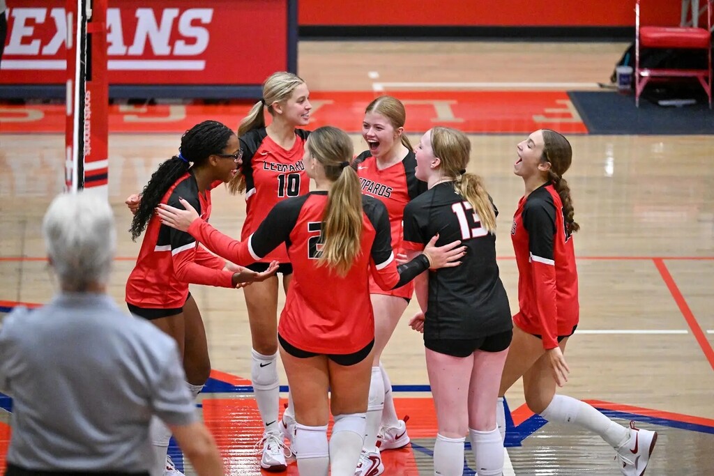 A team of female volleyball players in red uniforms with numbers huddle on a court. A referee in gray stands nearby.