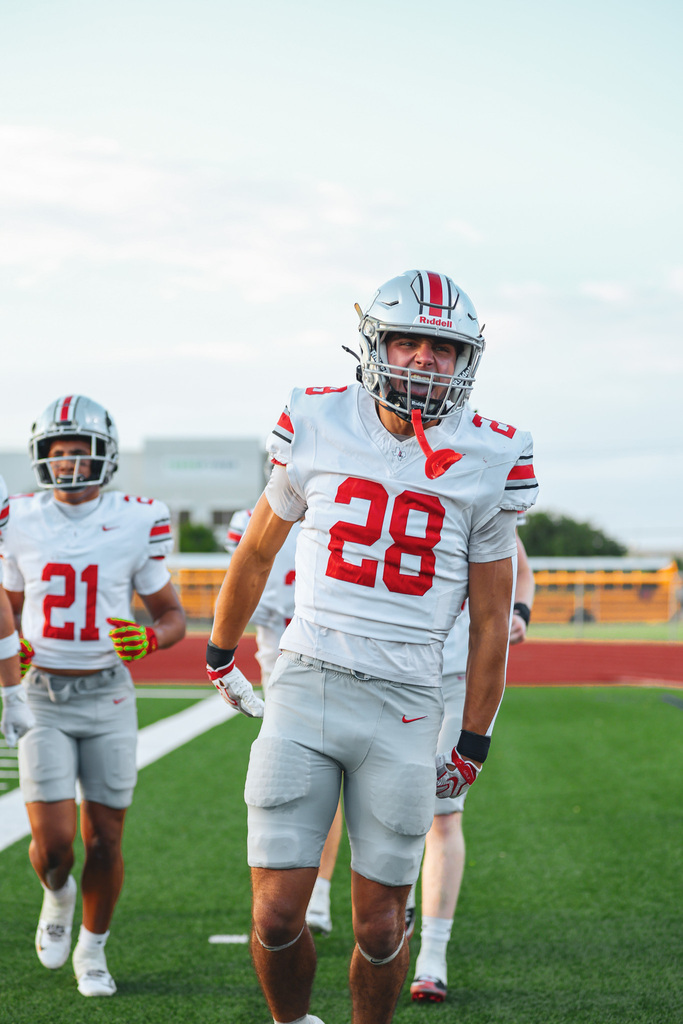 Football players wearing helmets and numbered jerseys running on a green field. One player wears a red and white jersey with the number 28.
