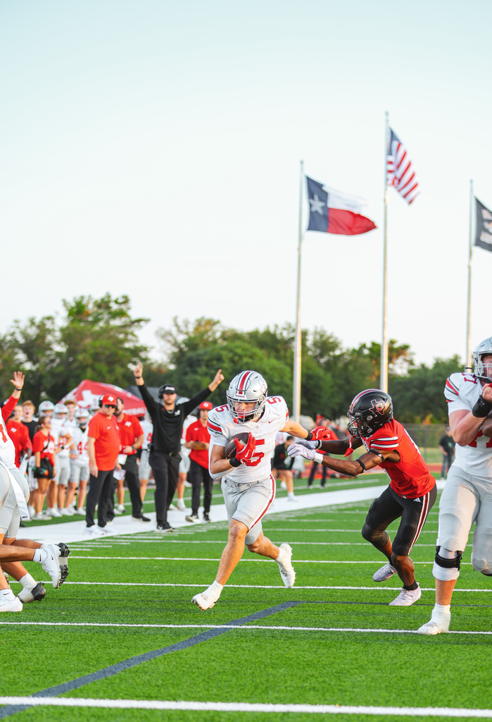 Players run on a football field. One player in a red uniform tackles another in white. Behind, flags, including a Texas flag, fly.