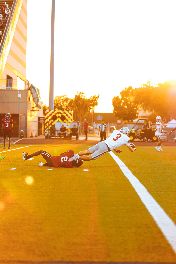 Players in jerseys fall on a field with a building, trees, and a setting sun in the background.