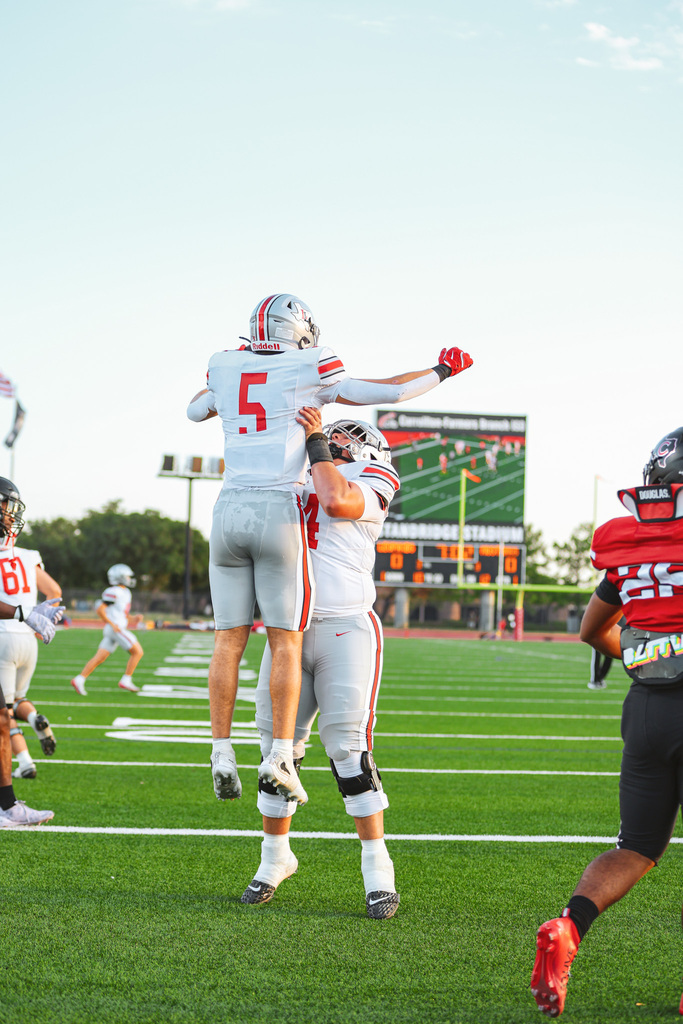 Two football players celebrate a touchdown. One player lifts the other on his shoulders. A scoreboard is in the background.