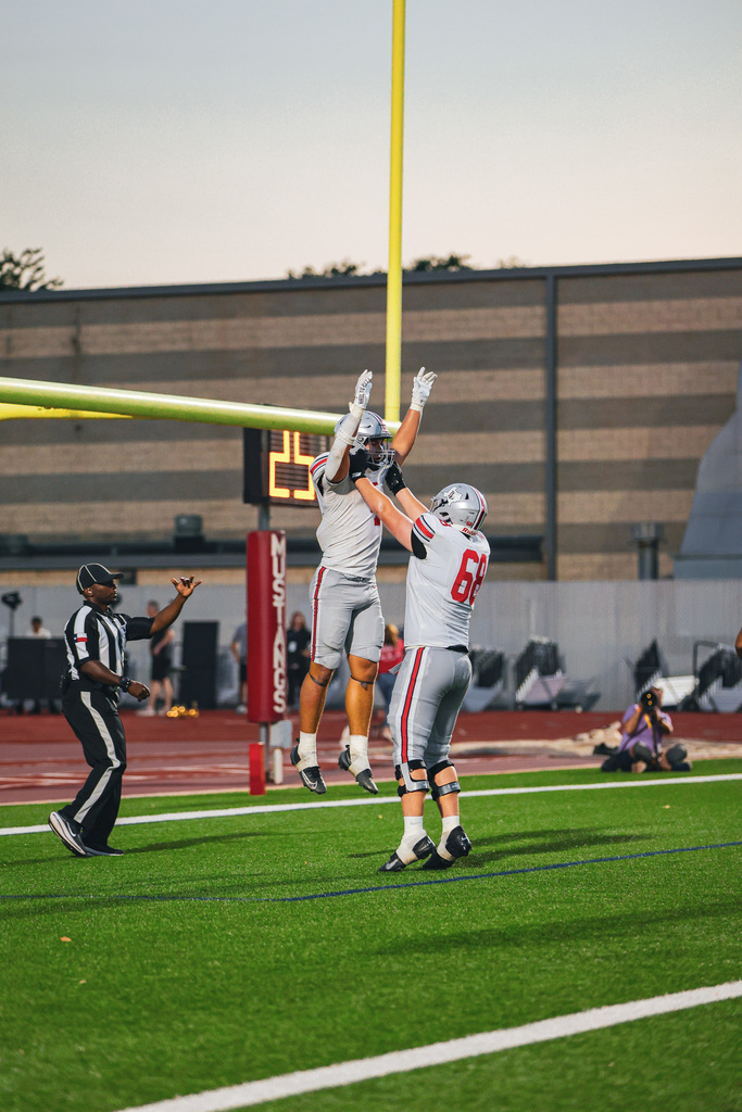 A football player jumps between two others to catch the ball. A referee watches from the sideline.