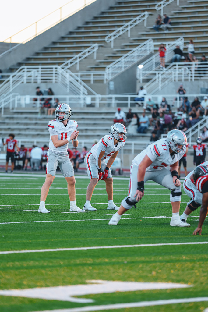 American football players in white jerseys and helmets on a field. The player in the middle holds a football, while others watch.