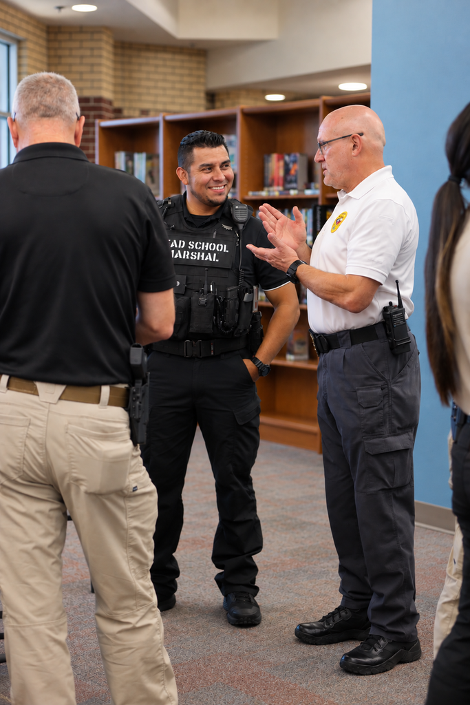 Three men in uniform stand in a hallway. One wears a black shirt, another a white shirt. Behind them are shelves with books.