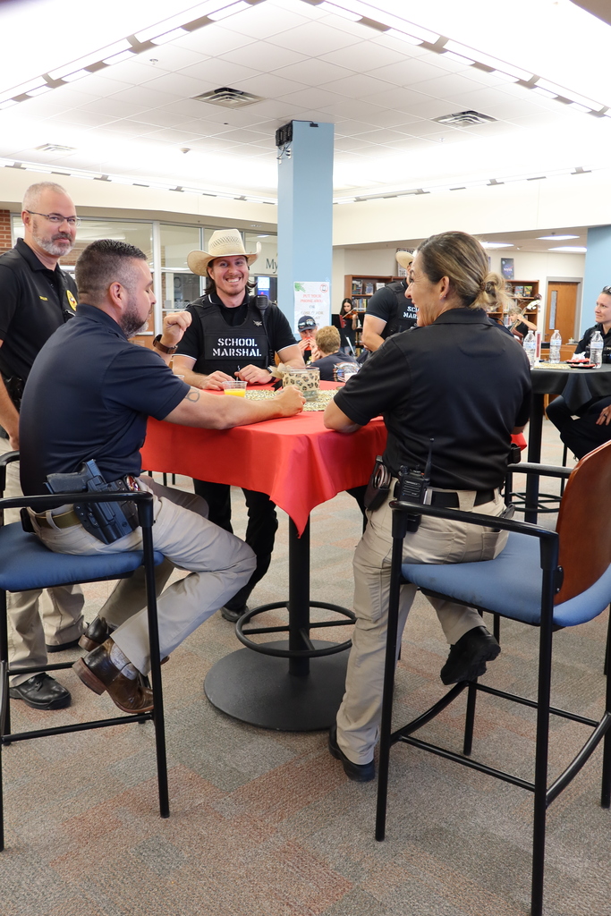 Six people in uniforms are seated at a table in a cafeteria. Three people sit on chairs and three stand.