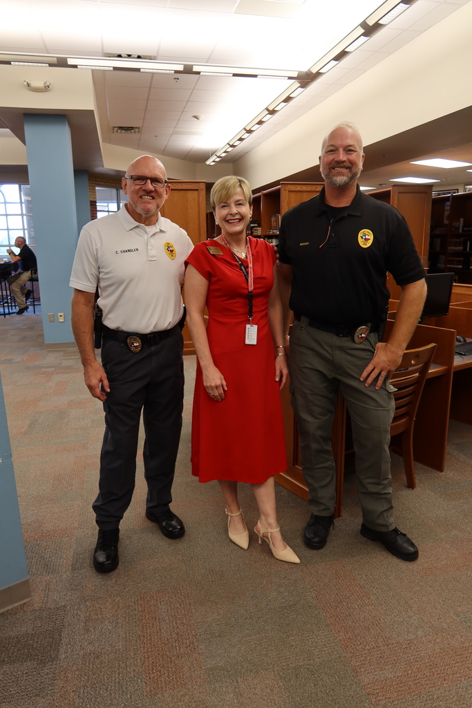 Three people stand in a room; two men wear uniforms and the woman wears a red dress.