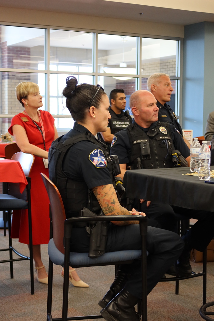 People sit in chairs around a table; one officer wears a vest. Behind them, glass windows and a door.