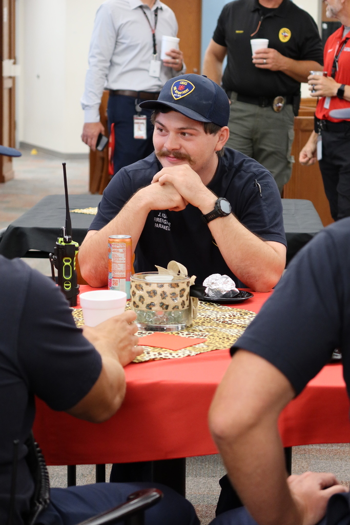 A man wearing a blue uniform and a hat sits at a table with food, talking to others.