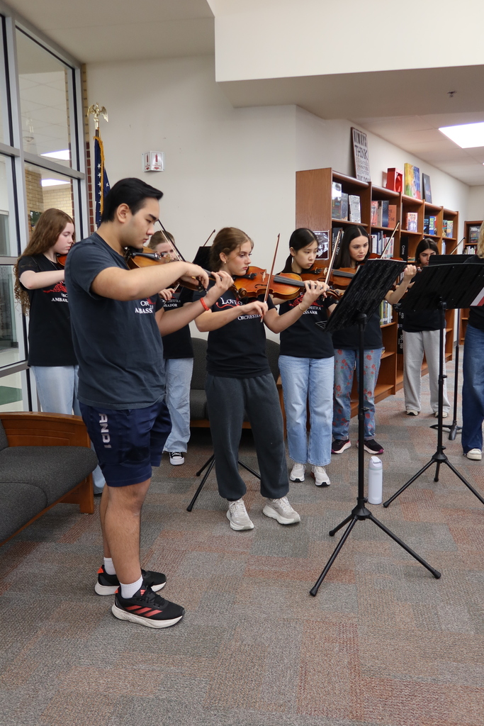 A group of young people play violins and stand in front of music stands. They are in a room with a white ceiling, bookshelves, and carpeted floor.
