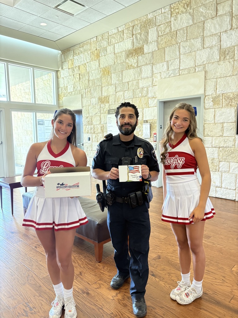 Two cheerleaders in red and white uniforms, one holding a box, stand next to a uniformed officer.