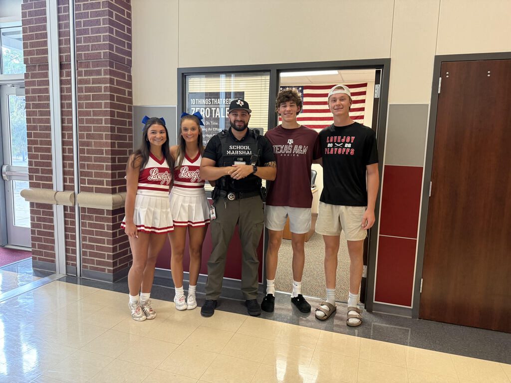 Five people stand together in a hallway. Two are cheerleaders, one in a police uniform, and two in casual clothes.
