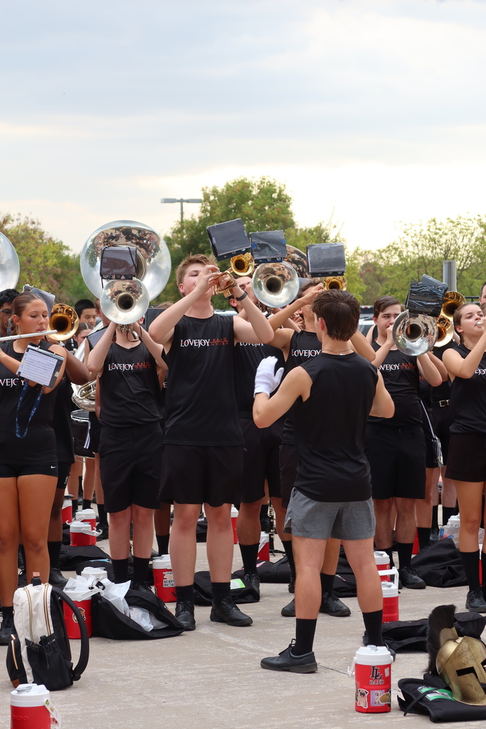 Band members in black uniforms stand in a row, holding instruments and standing around coolers on a paved surface.