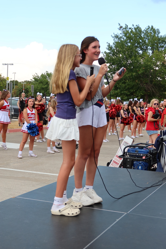 Two girls on stage with a microphone; one holds it, the other stands behind her. A crowd of cheerleaders in red and white uniforms is in the background.