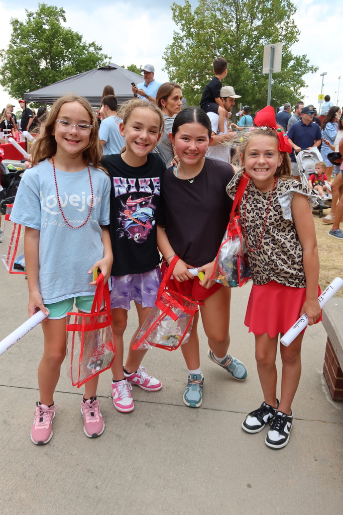 Four girls stand on a sidewalk, smiling, holding bags. Behind them, a busy area with people, trees, and a canopy.