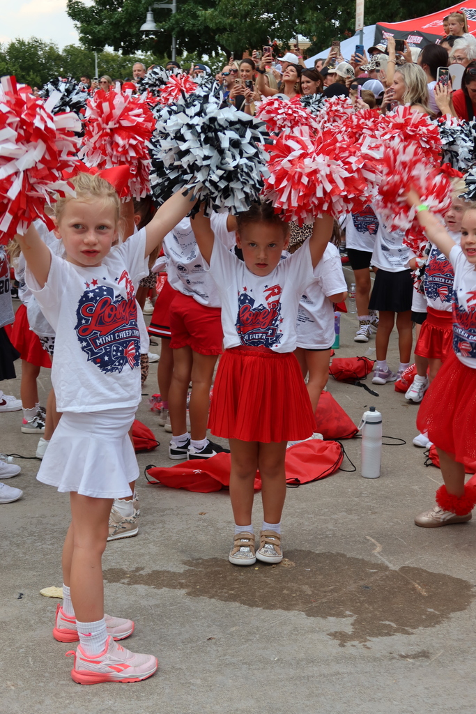 Cheerleaders in red and white uniforms hold up pom-poms; others watch in background. Concrete ground and streetlights in distance.