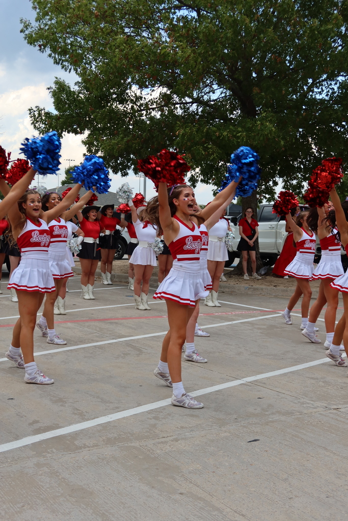 Cheerleaders in red and white outfits perform with blue and red pom-poms. Trees and a parking lot in the background.